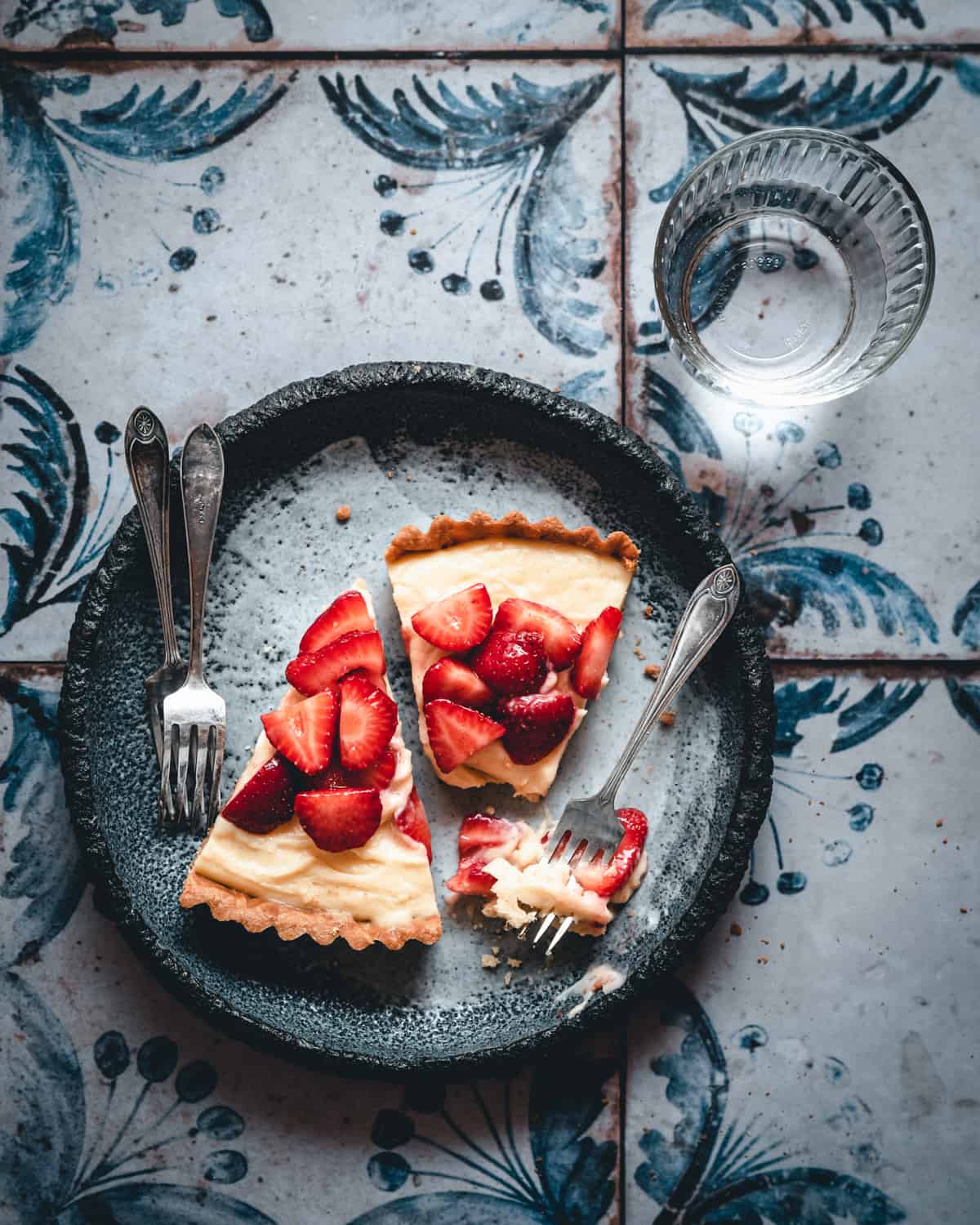 Two slices of Tarte aux Fraises on a plate with forks and glass of water.